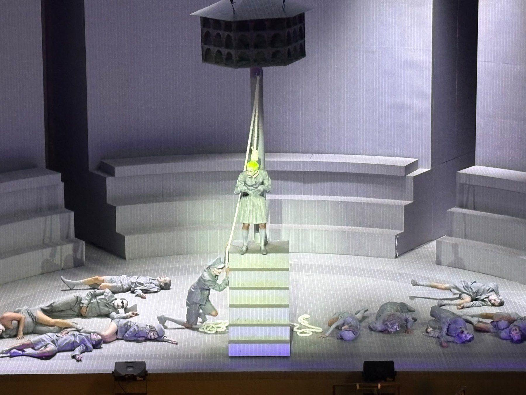Figure with green hair on a staircase, bodies lying on the grey stage